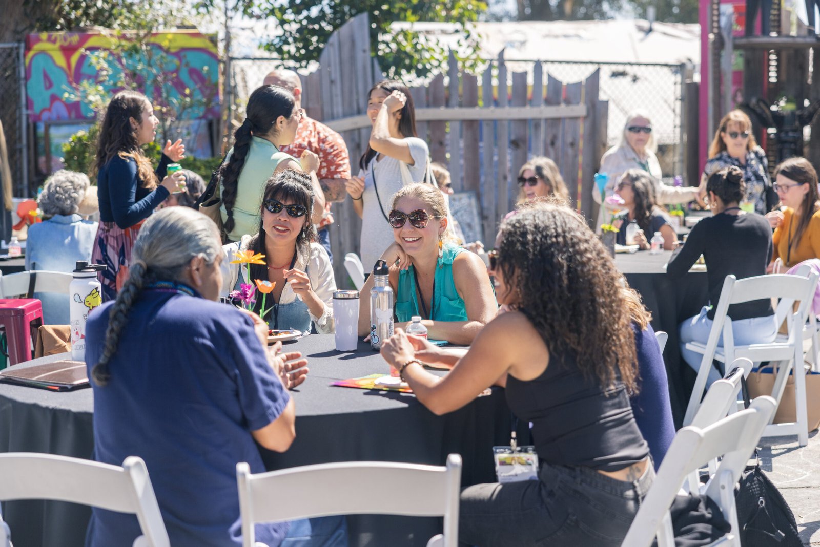 A group of people are seated at round tables covered in black tablecloths at an outdoor event. They are in conversation and smiling, with water bottles, flowers, and drinks on the tables. White folding chairs surround each table. A few attendees are standing, talking, or walking in the background. A colorful painted mural and wooden fence frame the background.