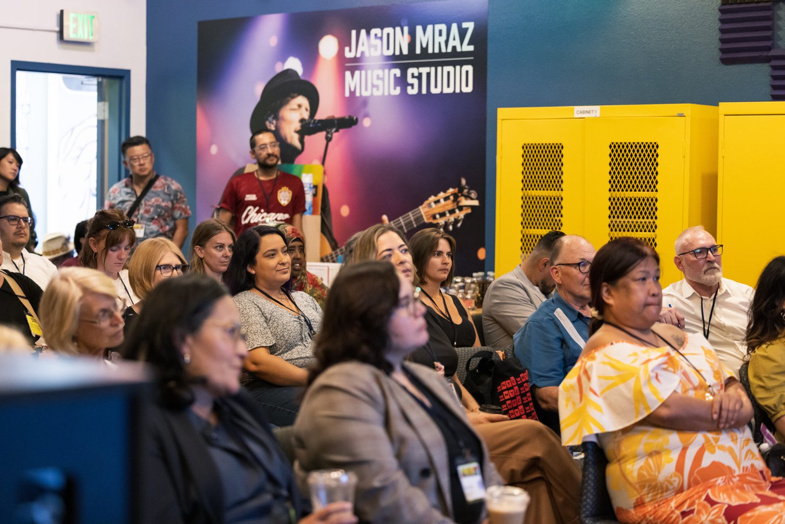 Audience members seated in a conference room with a large "Jason Mraz Music Studio" banner in the background, yellow cabinets to the right.