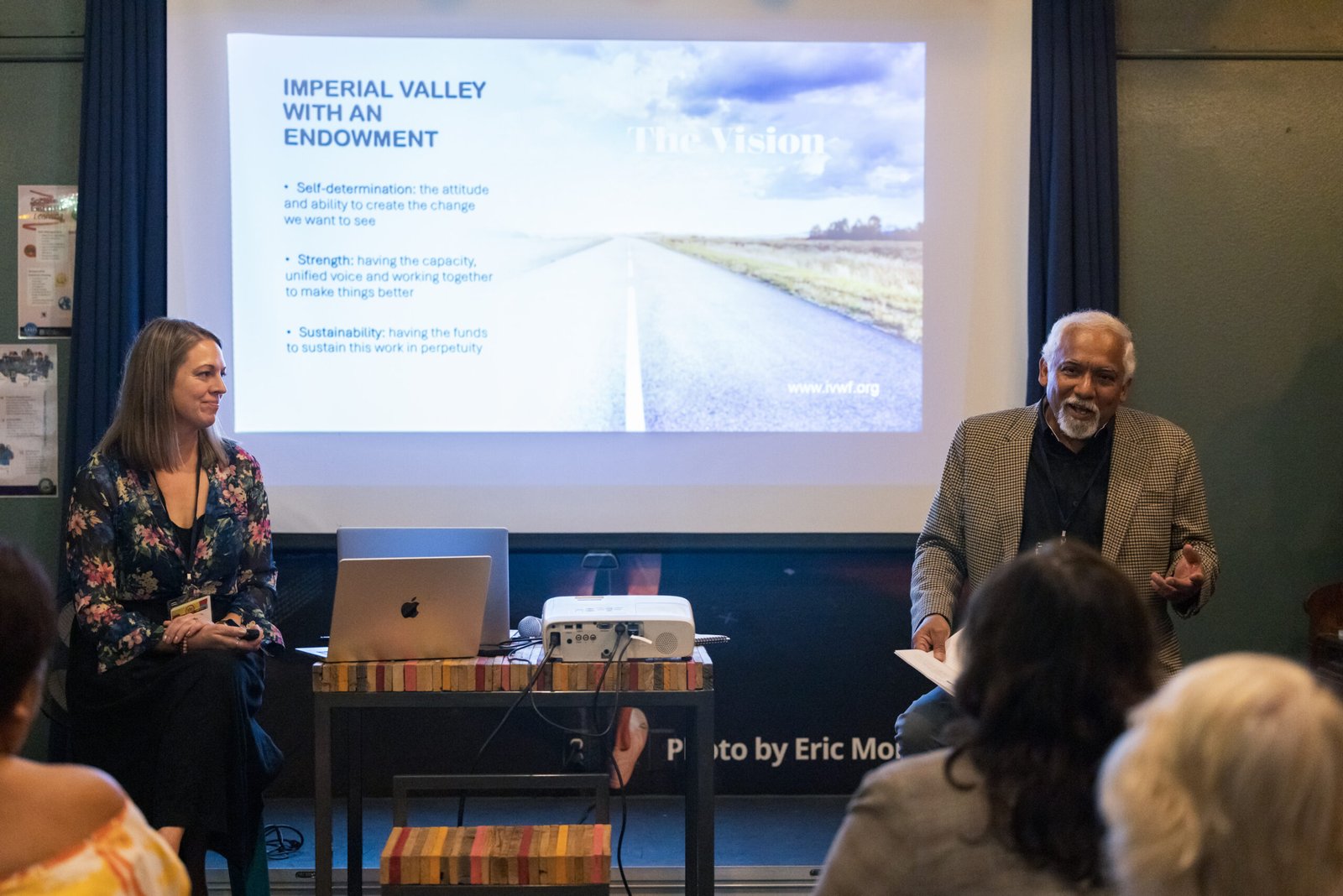 A man and a woman sitting on a stage in front of a presentation screen, addressing an audience. The screen displays the text "IMPERIAL VALLEY WITH AN ENDOWMENT" with bullet points detailing the organization's goals, and an image of a road. The woman on the left is wearing a floral blouse and looking towards the right, while the man on the right, wearing a patterned jacket, is gesturing with his hands. A laptop and projector are on a table in front of them. The audience is blurred in the foreground.