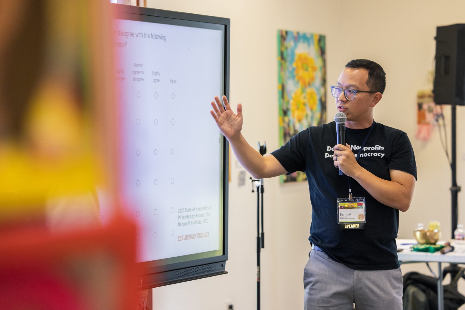 A man with glasses and a microphone giving a presentation, gesturing towards a large screen displaying a survey about nonprofits and philanthropy. He is wearing a black t-shirt that says "Nonprofits Democracy" and a lanyard with a "Speaker" badge.