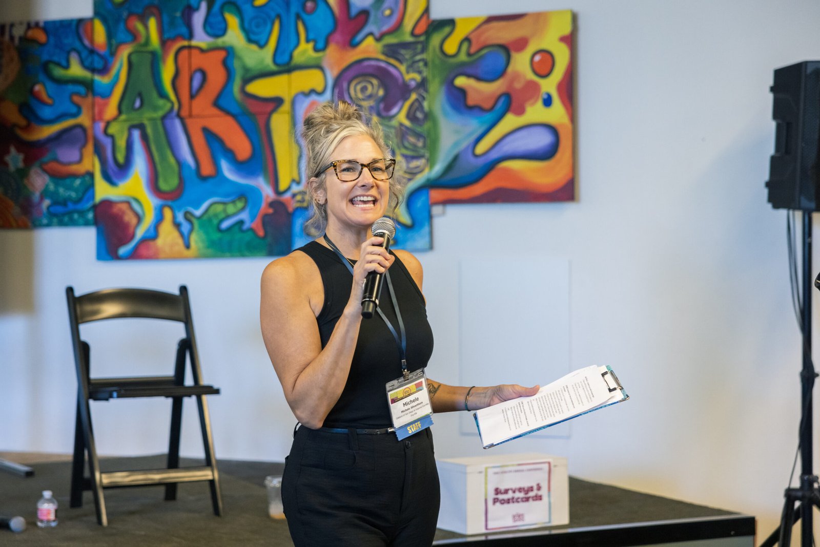 A woman with glasses and a microphone speaks at a conference, holding a clipboard, standing in front of abstract colorful artwork with "ART" visible on it. A black chair, a speaker stand, and a box labeled "Surveys & Postcards" are also visible.