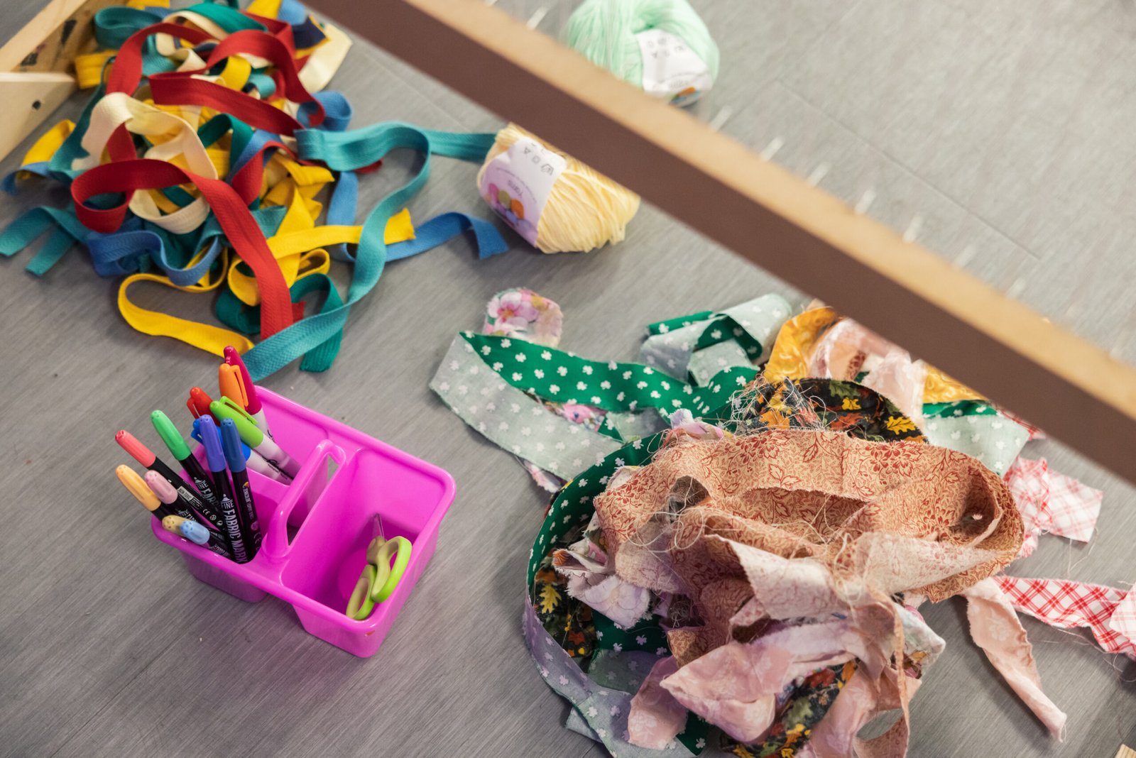 A collection of crafting supplies on a gray surface, including colorful strips of fabric and zipper tape, yarn, fabric scraps, fabric markers in a pink caddy, and scissors, viewed from an upward angle.