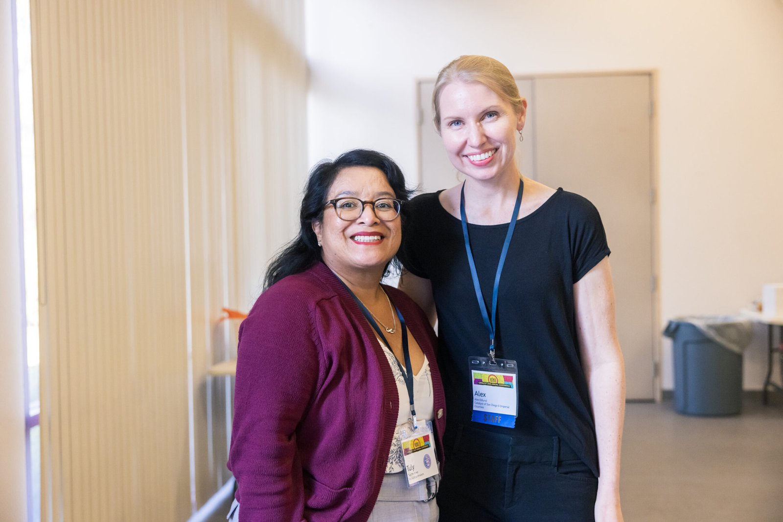 Two smiling women posing for a picture at a conference. The woman on the left, wearing glasses, a burgundy cardigan, and a name tag that says "Tuly," smiles widely. The woman on the right, with blonde hair and a black top and pants, has a name tag that reads "Alex." They are standing in a well-lit hallway with vertical blinds on one side.