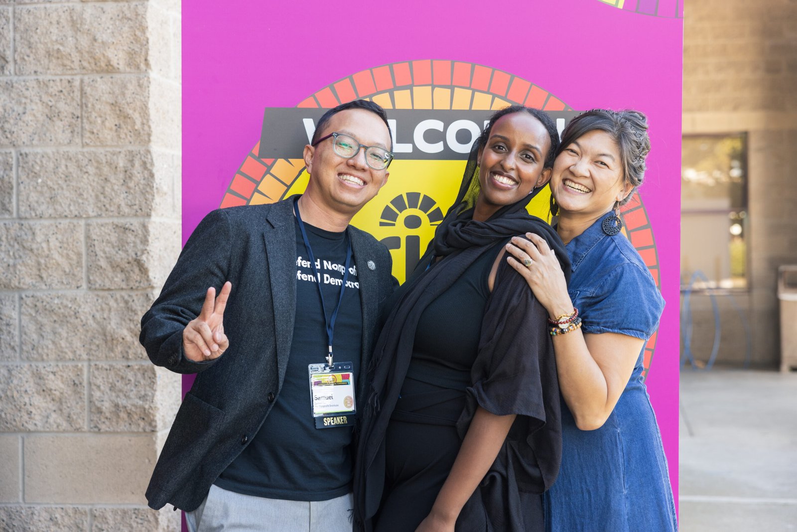 Three diverse people smiling and posing for a photo in front of a colorful "Welcome" sign, a man giving the peace sign and wearing a speaker badge.
