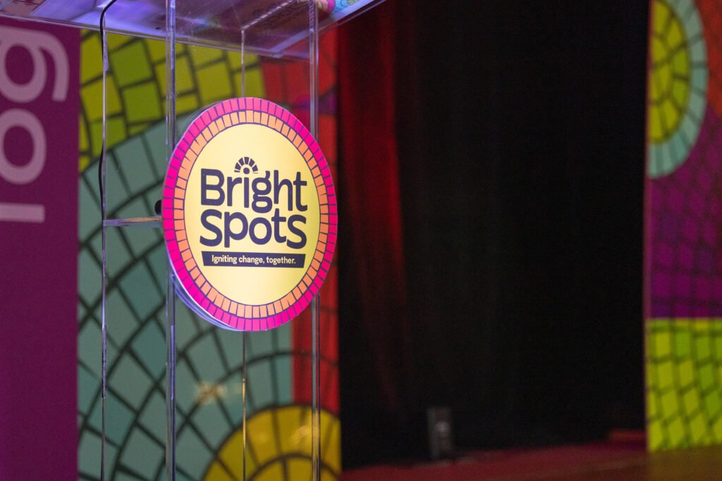 A circular sign for "Bright Spots" with a yellow center, orange and pink trim, and black text against a blurred colorful background. The sign is inside a clear glass encasing. The background includes purple, green, and red panels.