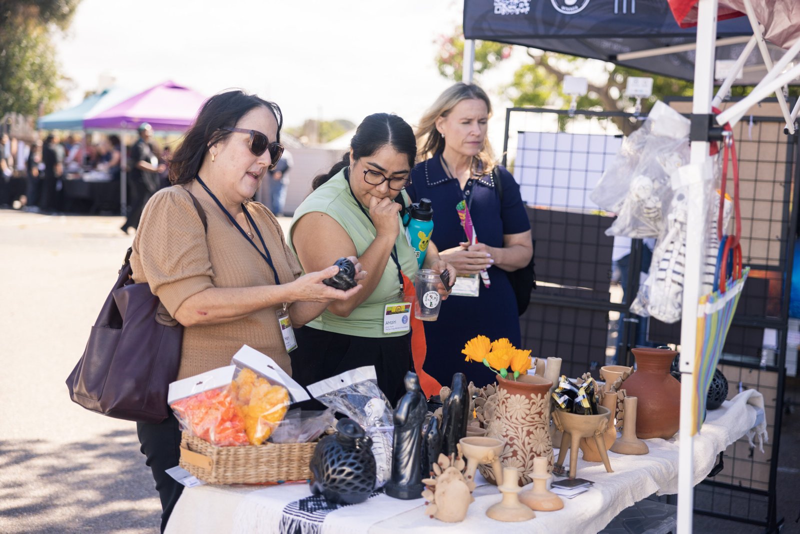 Three women examining handcrafted items at an outdoor artisan market; one woman holds a small, black ceramic piece, while the others observe; a table in the foreground displays various pottery, ceramic figurines, and packaged treats; the background reveals additional vendor booths and attendees.