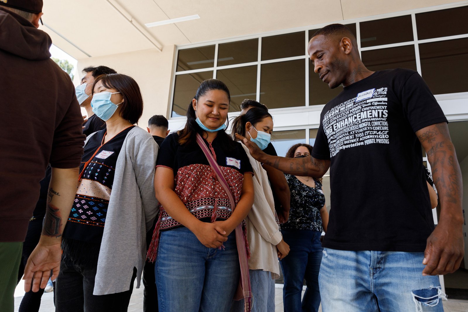 Community Safety Training 106 A group of people, most wearing face masks, standing in line and facing forward outside a building. A man in a black t-shirt with "ENHANCEMENTS" in large white text and smaller text with legal terms touching the shoulder of a woman wearing a mask and a patterned top. A name tag reads "Paw Say." Another name tag reads "Nao Kalsberg 1993." A name tag reads "Michael Camper."