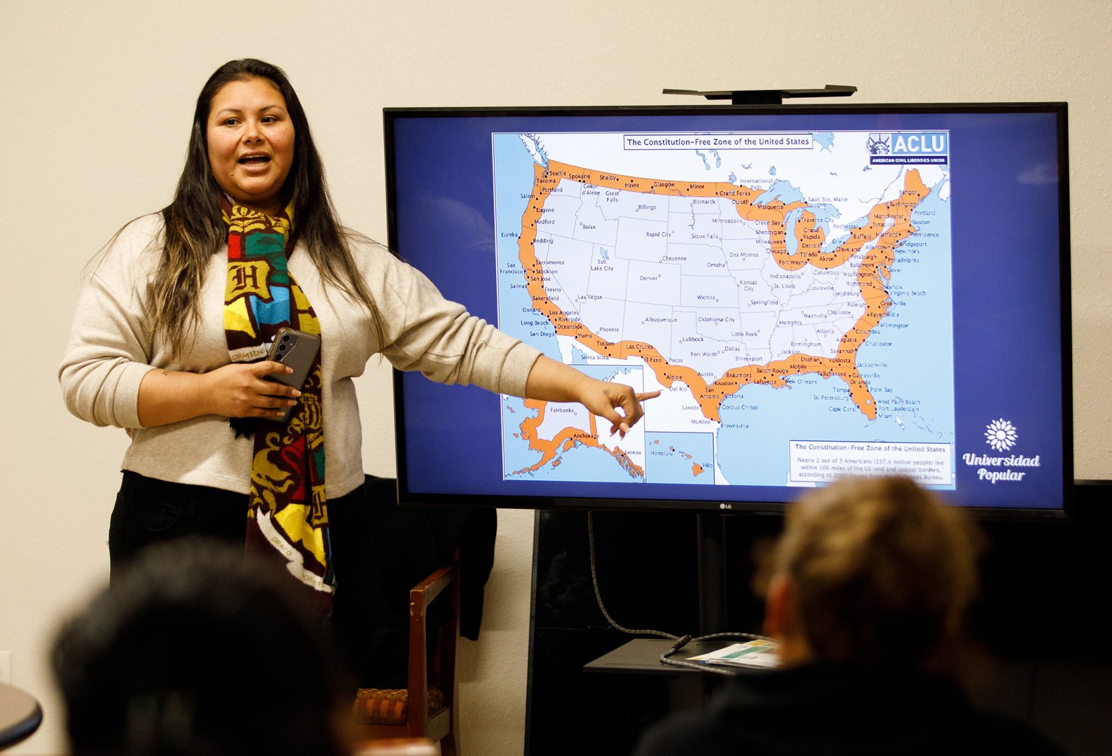 MigrantDayResourceFair 055 A woman with dark hair, wearing a patterned scarf with a "H" graphic, stands in front of a screen displaying a map of the United States with highlighted areas and the title "The Constitution-Free Zone of the United States," alongside the ACLU logo. The text on the screen includes city names, geographic markings, and the statement: "Nearly 2 out of 3 Americans (197.4 million people) live within 100 miles of the US land and coastal borders, according to 2007 figures from the Census Bureau." The screen also features the "Universidad Popular" logo. The woman is gesturing with her hand towards the map. Several blurred individuals are in the foreground, indicating an audience or group setting.