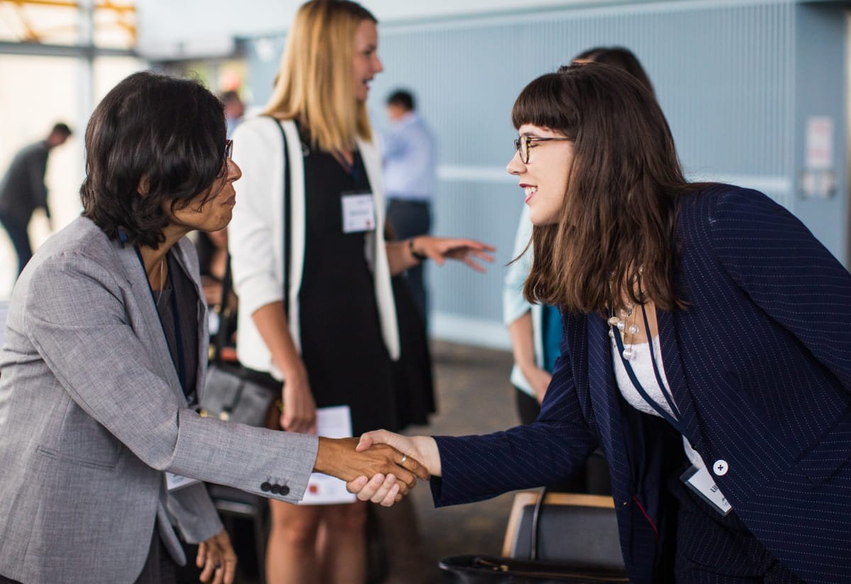 Image Preview Two women in business attire shaking hands during an event, other attendees visible in the background.