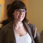Courage Young woman with brown hair and glasses smiling, wearing a grey blazer over a white lace top, and carrying a pink purse.