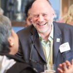 Impact A smiling, middle-aged bald man in a suit jacket and glasses hanging from his shirt, talking and gesturing at an event; a name tag reads "Tomas Herrera-Mishler."