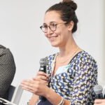 Learning Image Preview A smiling woman with glasses and a bun, wearing a patterned blue and white blouse, holding a microphone and gesturing during a panel discussion.