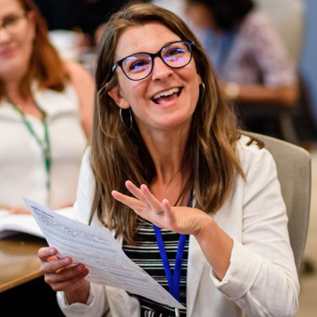 Philanthropy Ca 04 Woman with long brown hair wearing glasses, smiling and gesturing with her hands while holding a document during a conference or meeting.