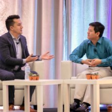 Philanthropy Ca 1 Two men in business attire seated at a table on a stage, engaged in a conversation, gesturing with hands, with potted succulents on the table, and a backdrop of vertical fabric panels.
