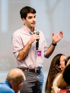 Philanthropy Ca 3 A young man with dark hair, wearing a light blue checked shirt and belt, holding a microphone, gesturing with his right hand as he speaks to an audience; a purple name tag is on his chest.